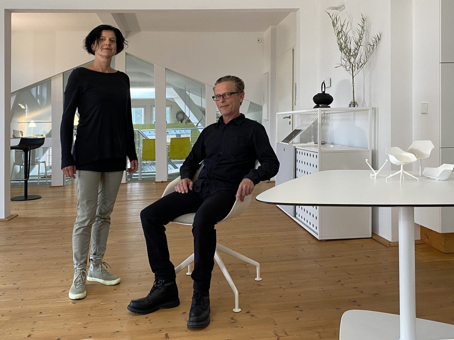 The two people are in a bright, modern office or studio space. The man is sitting on a Yonda, recognizable by its distinctive shell-shaped seat and delicate base. There are also Yonda design models on the table in the foreground.