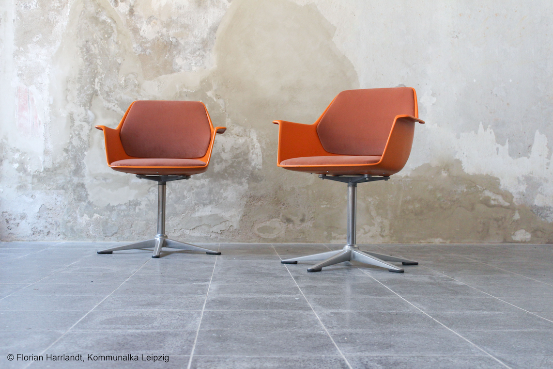 Here are two chairs in a warm orange hue. The chairs combine clean lines with a sturdy four-star metal base. Against the rough concrete wall, an intriguing contrast emerges between modern design and the industrial setting.