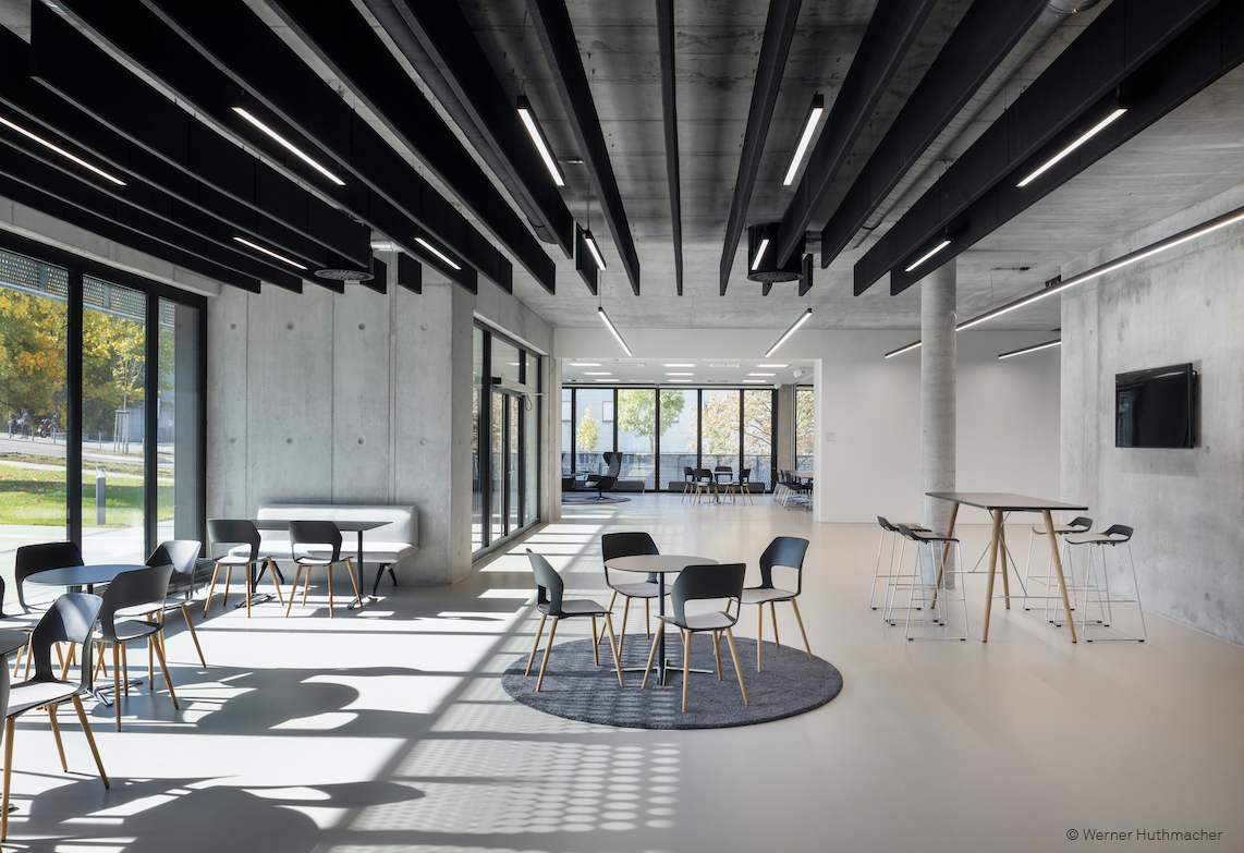 The bright interior features an open lounge and break area with various seating arrangements and tall bar tables. The black shell chairs with wooden legs closely resemble the Occo chairs, which are frequently used in modern communication areas. The space feels inviting and versatile.