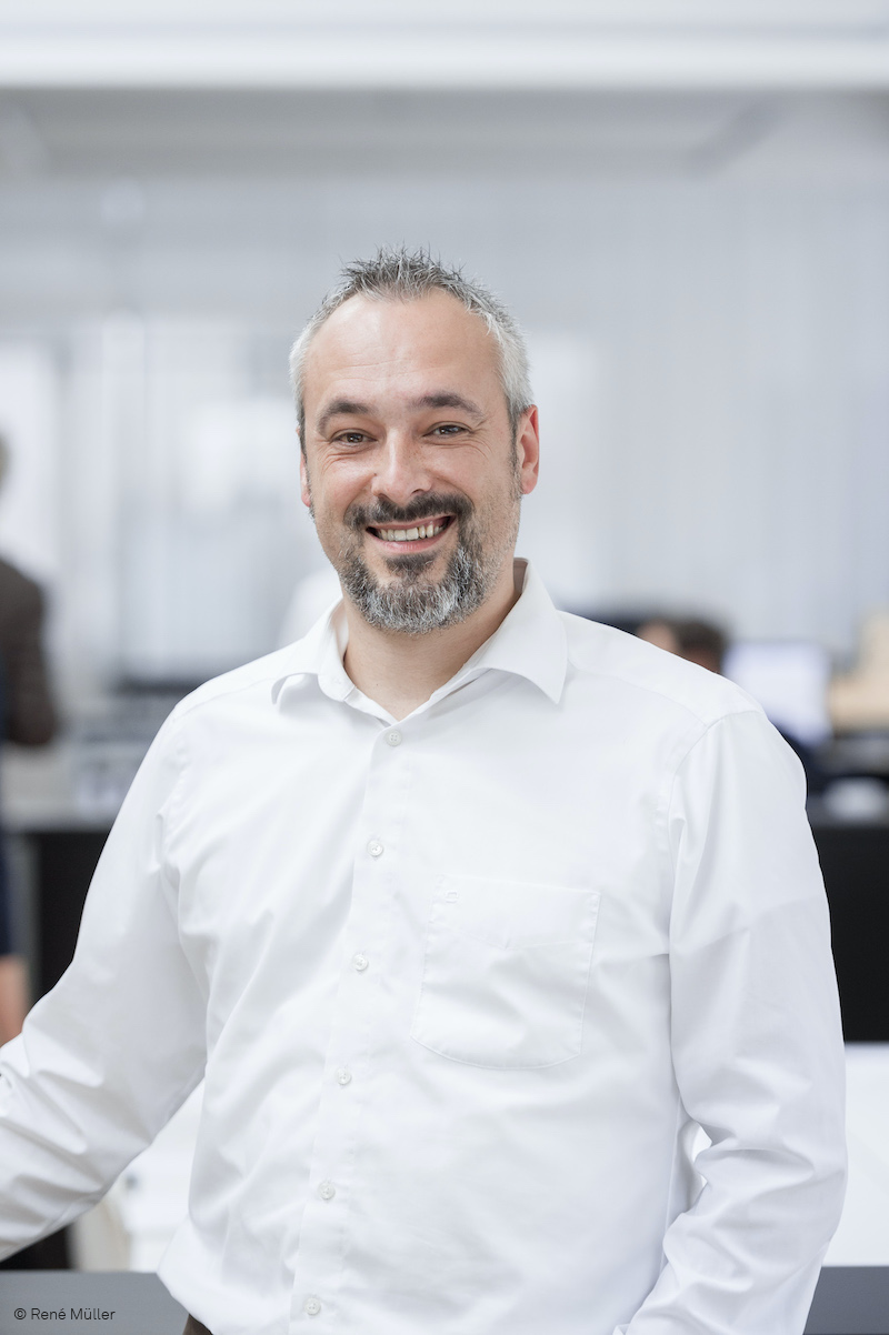 The photo shows a man smiling warmly in a white shirt in a bright, modern office setting. The background is blurred, drawing the viewer’s attention entirely to the person.