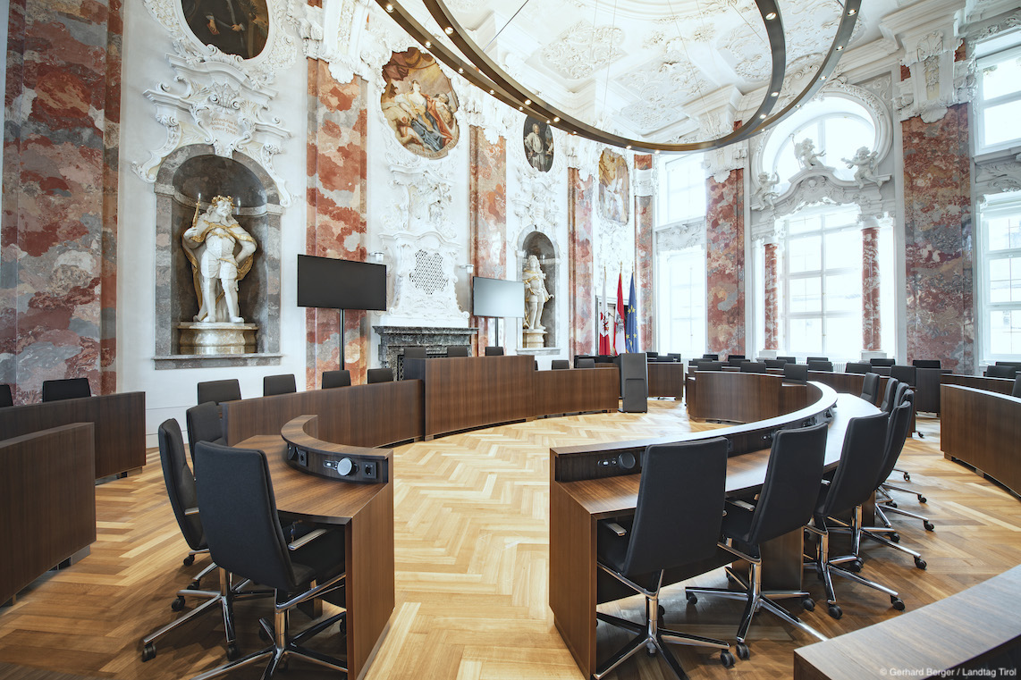 Many black Sola conference chairs are arranged in circular rows. They are set up in a large hall featuring extravagant wall paintings.