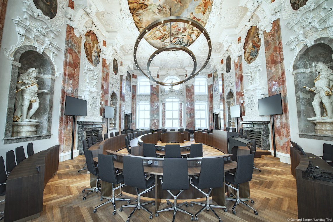 Many black Sola conference chairs are arranged in circular rows. They are set up in a large hall featuring extravagant wall paintings.
