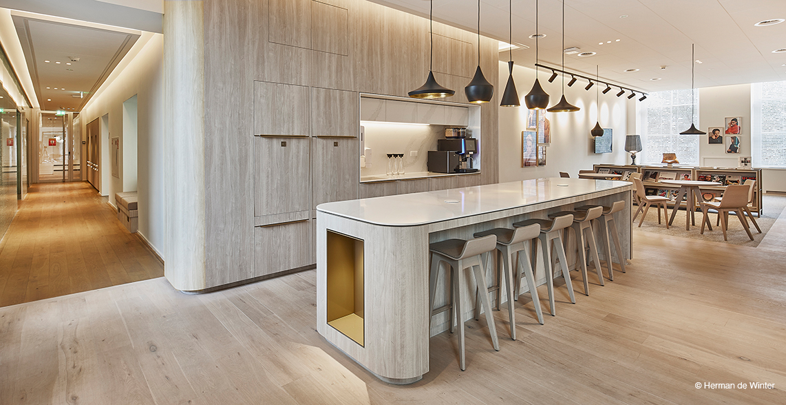 The space features a stylish lounge or common area with a light wood finish, a long kitchen island, and modern stools. In the back, there are several lightweight, modern chairs. The room feels inviting and conducive to conversation, making it ideal for breaks or informal meetings.