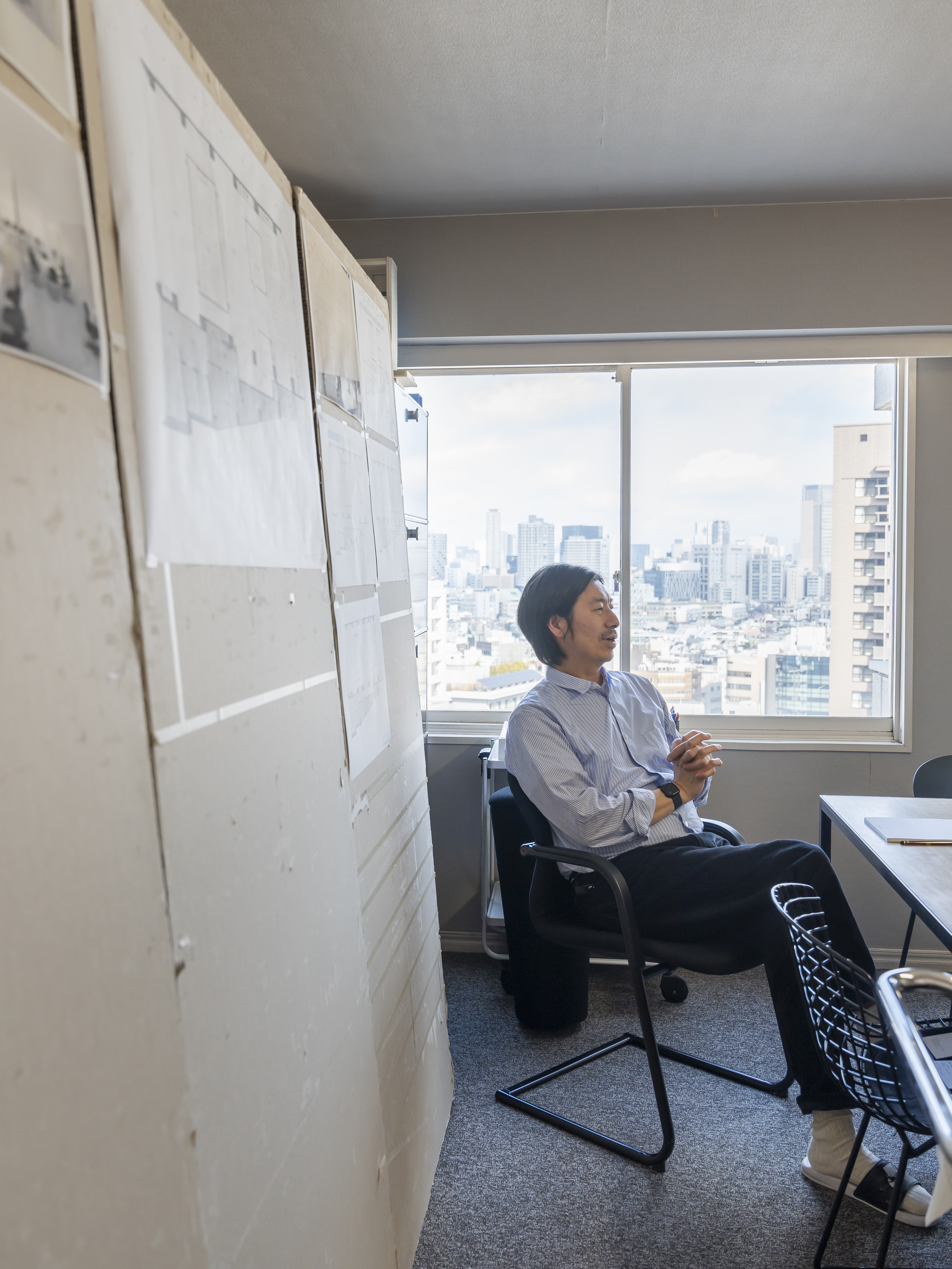 A man is sitting in an office in front of a large window overlooking a skyline. The setting feels creative, personal, and slightly improvised.