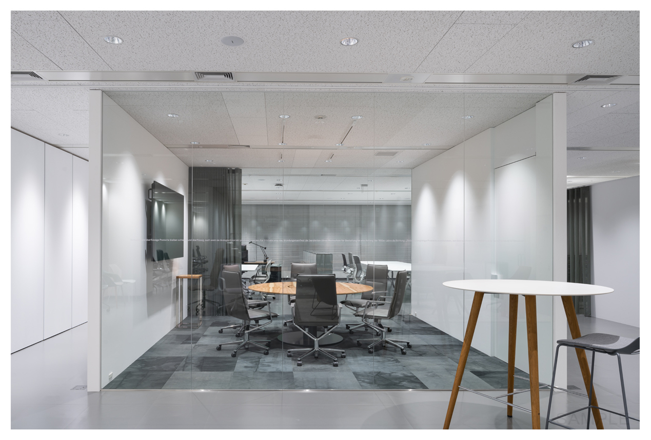 The room is a bright, modern meeting room featuring a round wooden table and several sleek gray Sola swivel chairs. In the foreground on the right, you can see a tall Occo table with wooden legs and a white tabletop.