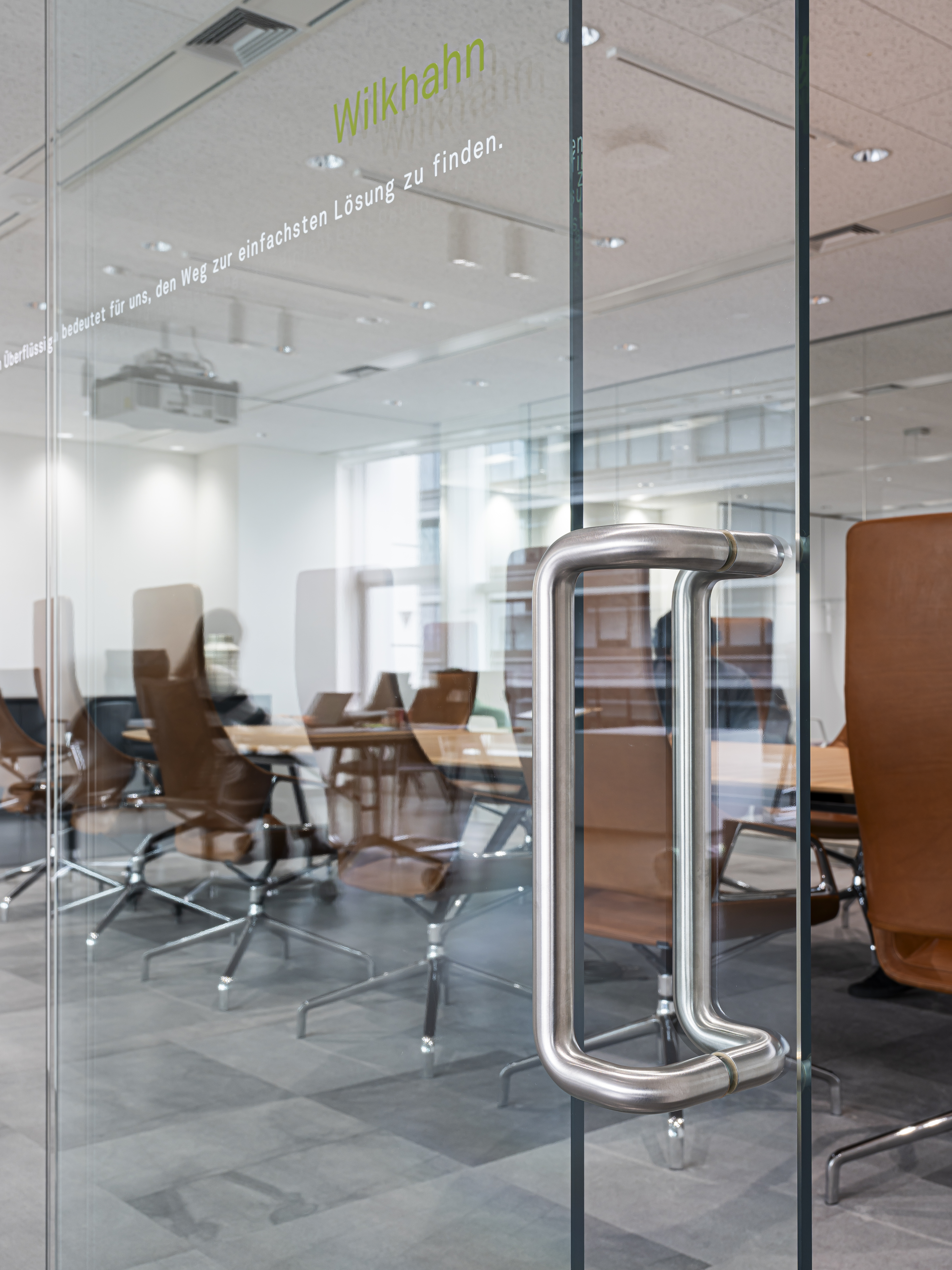 The photo shows a close-up of the glass door to the conference room, with the “Wilkhahn” logo clearly visible. Behind it, you can once again see the Graph conference chairs with brown leather upholstery and high backrests. These conference chairs define the space. The image highlights the high-quality, brand-focused entrance area.